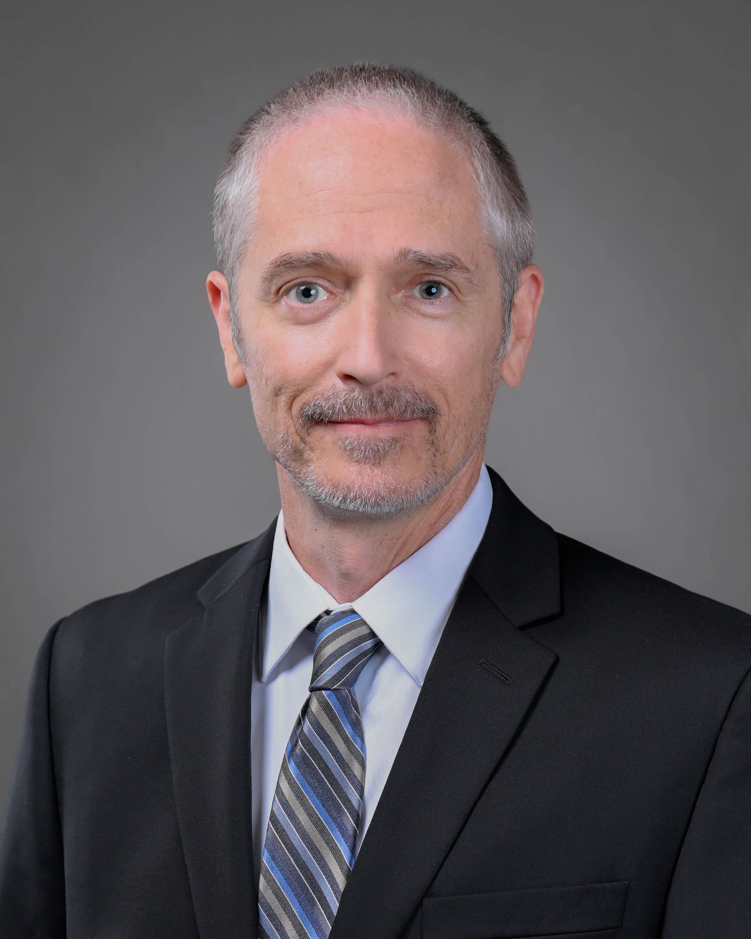grey and brown haired man with a short goatee smiling in a dark suit with a striped tie.